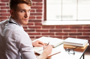 Handsome young man in shirt and tie writing something in note pad and looking over shoulder while sitting at his working place
